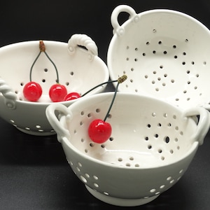 Può includere: Tre scolapasta in ceramica bianca con manici, due sono impilati e uno è sul tavolo. Lo scolapasta sul tavolo ha tre ciliegie rosse all'interno.