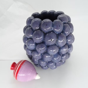 Pode incluir: Um vaso de cerâmica com um exterior texturizado roxo escuro, assemelhando-se a um cacho de uvas. Ao lado, um pequeno pião pintado com listras vermelhas e brancas e uma ponta branca. O vaso é um item decorativo único.