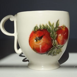 May include: White ceramic mug with a hand-painted design of two red pomegranates with green leaves.