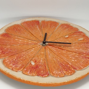 Puede incluir: Un reloj de pared de cerámica con forma de rodaja de naranja. La esfera del reloj es naranja con un borde blanco y tiene agujas negras.