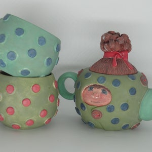 May include: A green teapot with a woman's face and a red bow on top. The teapot is decorated with blue and red polka dots. Two green cups with polka dots are stacked next to the teapot.