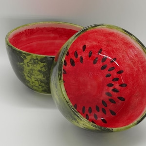 May include: Two ceramic bowls shaped like watermelon halves. The interior is bright red with black seed details, while the exterior is green with a textured pattern. The bowls are designed to resemble the fruit.