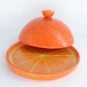 Könnte beinhalten: Orangefarbene Keramik-Schale mit einem Deckel in Form einer Orange. Die Schale ist eine Orangenscheibe und der Deckel ist eine ganze Orange.