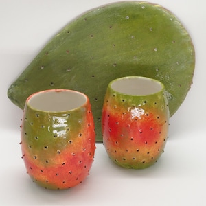 Op de afbeelding: Twee keramische bekers met een groen, oranje en rood cactusontwerp. De bekers staan op een wit oppervlak.