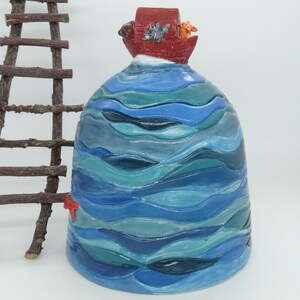 May include: A ceramic sculpture depicting Noah's Ark. The red ark sits on a base of blue and teal waves, with animal figures on top. A small wooden ladder is to the left, and a red starfish is on the side.