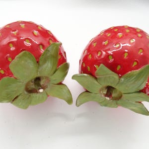 Peut inclure: Deux fraises en céramique avec des surfaces rouges brillantes et des dessus verts et feuillus. Les fraises ont de petites graines jaunes et sont posées sur un fond blanc. Idéal pour la décoration de cuisine ou de salle à manger.