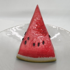 May include: A slice of watermelon with a red interior and black seeds, resting on a white plate. The rind is a light green color with a white border. The watermelon slice is a triangular shape.