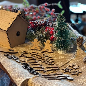 Pu&ograve; includere: Un kit di casetta di pan di zenzero in legno marrone con un pino innevato, due omini di pan di zenzero e vari ritagli di legno per decorare. Il kit include stelle, fiocchi di neve, foglie e altre forme festive.