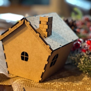 Pu&ograve; includere: Una piccola casa di legno marrone con un camino e un tetto nero. La casa ha due finestre ed &egrave; decorata con dettagli intricati.