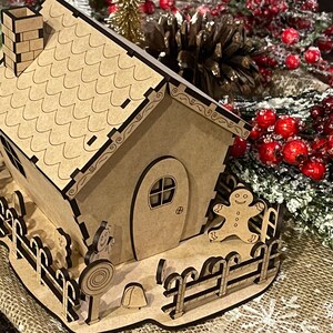 Pu&ograve; includere: Una casetta di pan di zenzero in legno con un tetto marrone, un camino e un omino di pan di zenzero in piedi all'esterno. La casa ha una piccola recinzione e una canna di zucchero sul terreno.
