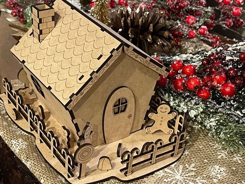 Pu&ograve; includere: Una casetta di pan di zenzero in legno con un tetto marrone, un camino e un omino di pan di zenzero in piedi all'esterno. La casa ha una piccola recinzione e una canna di zucchero sul terreno.