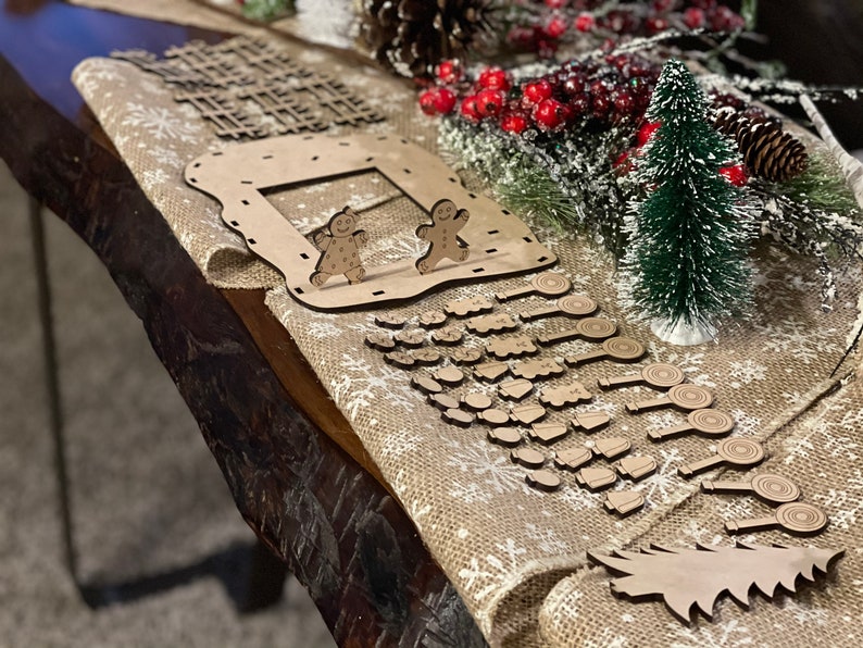 Pu&ograve; includere: Un calendario dell'avvento natalizio in legno con una casetta di pan di zenzero, omini di pan di zenzero e un albero di Natale. Il calendario ha 24 porte numerate, ciascuna con un piccolo ornamento in legno dietro. Gli ornamenti includono bastoncini di zucchero, campane, stelle e altre forme festive.