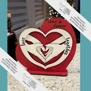 Puede incluir: Un recorte de madera en forma de corazón rojo con un interior blanco. El recorte presenta dos manos formando una forma de corazón. El texto "love" está en el lado izquierdo y "together" en el lado derecho. Los nombres "Amy & Trever" están en la parte superior del recorte.