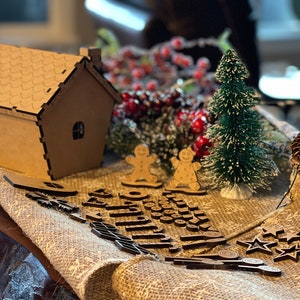Pu&ograve; includere: Una casetta di pan di zenzero in cartone marrone con un tetto bianco glassato, due omini di pan di zenzero, un piccolo albero di Natale verde e una variet&agrave; di decorazioni natalizie in legno, tra cui stelle, fiocchi di neve e una ghirlanda.