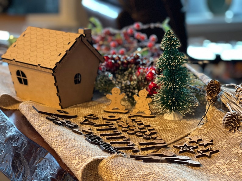 Pu&ograve; includere: Una casetta di pan di zenzero in cartone marrone con un tetto bianco glassato, due omini di pan di zenzero, un piccolo albero di Natale verde e una variet&agrave; di decorazioni natalizie in legno, tra cui stelle, fiocchi di neve e una ghirlanda.