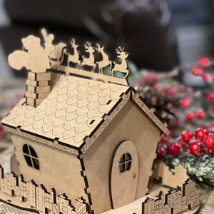 Pu&ograve; includere: Una casa di Natale in legno con un camino, un tetto con un design a festoni e una recinzione con regali. Babbo Natale &egrave; sul tetto con le renne.