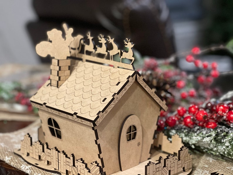 Pu&ograve; includere: Una casa di Natale in legno con un camino, un tetto con un design a festoni e una recinzione con regali. Babbo Natale &egrave; sul tetto con le renne.