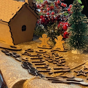 May include: A wooden gingerbread house kit with a small Christmas tree, two gingerbread people, and a variety of wooden cutouts, including stars, snowflakes, and swirls.
