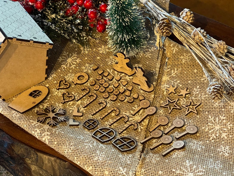 May include: A wooden gingerbread house kit with various cut-out pieces, including windows, stars, candy canes, and a snowman. The pieces are arranged on a burlap surface with a small pine tree and red berries.