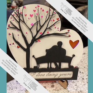 Könnte beinhalten: Ein Holzausschnitt eines Paares, das auf einer Bank unter einem Baum mit Herzen in den Ästen sitzt. Der Ausschnitt hat die Form eines Herzens und trägt den Text "Ich liebe es, dein zu sein" am unteren Rand. Der Ausschnitt ist für die Verwendung mit einer Glowforge konzipiert.