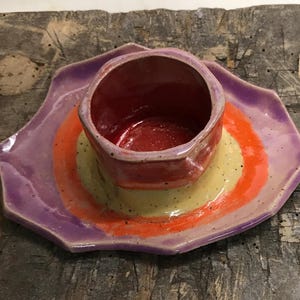 Könnte beinhalten: Handgefertigtes Keramik-Schalen- und Teller-Set. Die Schale ist tiefrot und steht auf einem Teller mit konzentrischen Kreisen in Gelb, Orange und Lila. Das Set steht auf einer rustikalen Holzoberfläche.