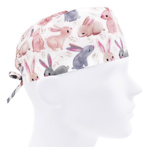 Op de afbeelding: Een witte scrub-muts met een roze en grijs konijnenpatroon. De muts heeft een strik aan de achterkant.