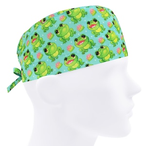Pode incluir: Um gorro cirúrgico azul turquesa com um padrão repetido de sapos verdes e lírios de água rosa. O gorro tem um laço na parte de trás.