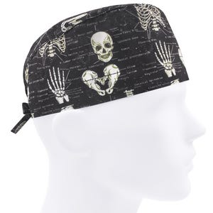 Op de afbeelding: Zwarte en witte scrub-muts met schedel- en bottenprint en anatomische labels. De muts heeft een schedel, botten en labels voor verschillende botten in het menselijk lichaam.