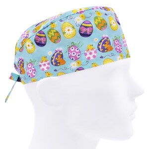 Op de afbeelding: Een lichtblauwe operatiemuts met een herhalend patroon van paaseieren, kuikens en bloemen. De eieren zijn versierd met strikken, stippen en bloemmotieven in verschillende kleuren. De muts heeft een striksluiting.