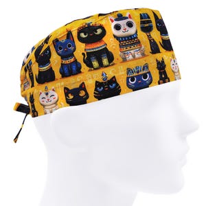 Puede incluir: Un gorro quirúrgico amarillo con un estampado repetido de gatos estilizados en negro, azul y blanco con diseños de inspiración egipcia. El gorro tiene un cierre de lazo y se lleva en una cabeza de maniquí.