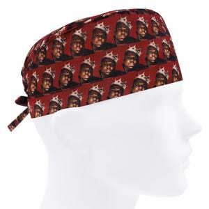 Könnte beinhalten: Eine weinrote OP-Haube mit einem sich wiederholenden Muster eines gekrönten Mannes auf rotem Hintergrund. Die Haube hat einen Kordelverschluss und wird auf einem weißen Schaufensterkopf präsentiert.