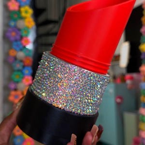 May include: A cylindrical container shaped like a lipstick tube with a bright red top section, a middle band covered in dense, shimmering silver rhinestones, and a solid black base. The object features a slanted top edge and a smooth finish.