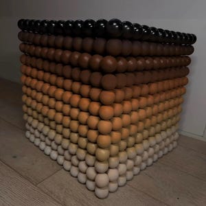 May include: A cube-shaped decorative object constructed from wooden spheres. The spheres transition in color from black at the top to shades of brown, orange, and finally, white at the base. The cube is placed on a wooden floor.