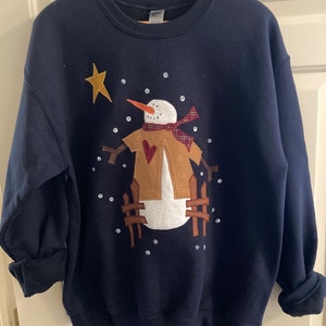 May include: A navy blue sweatshirt with a snowman applique. The snowman is wearing a brown hat, a red and black plaid scarf, and a brown coat with a red heart. The snowman is surrounded by white snowflakes.