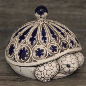 Puede incluir: Un joyero de cerámica con forma de tortuga y tapa extraíble. El caparazón presenta un patrón de arco gótico en relieve de color crema con inserciones esmaltadas en azul profundo. La cabeza y las patas tienen una textura escamosa en acabado mate.