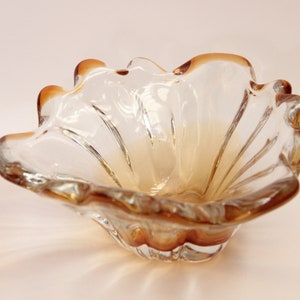 May include: A clear glass bowl with a shell-like shape and a wavy, scalloped rim. The glass features a gradient of amber-colored tinting that is concentrated along the edges and deepens toward the base, with vertical ridges along the interior.