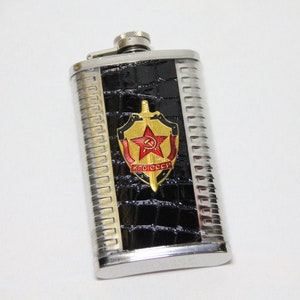 May include: A silver flask with a black crocodile-embossed design. The flask features a gold and red emblem with a sword and the text "CCCP".