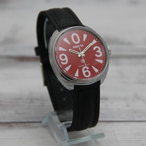 May include: A wristwatch with a red face and white numerals, including the number 3, 6, and 9. The watch has a silver-colored case and a black leather band. The word "RAKETA" is printed on the watch face.