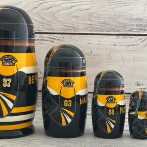 May include: A set of five black, yellow, and white nesting dolls. Each doll features a hockey jersey design with numbers and team logos. The largest doll has the number 37 and the word "BER" on it.