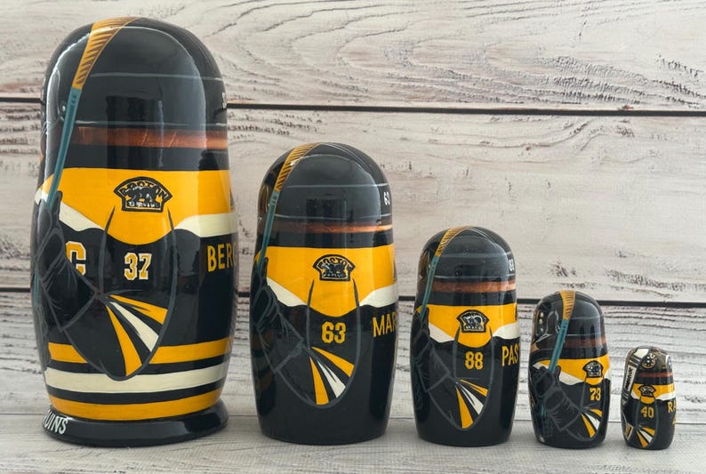 May include: A set of five black, yellow, and white nesting dolls. Each doll features a hockey jersey design with numbers and team logos. The largest doll has the number 37 and the word "BER" on it.