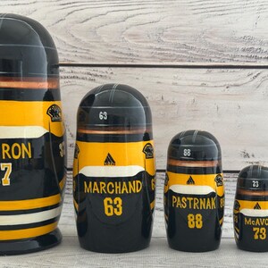 May include: A set of five black, yellow, and white nesting dolls. Each doll features a hockey helmet design and the names and numbers of hockey players: Bergeron 37, Marchand 63, Pastrnak 88, McAvoy 73, and Rask 40.