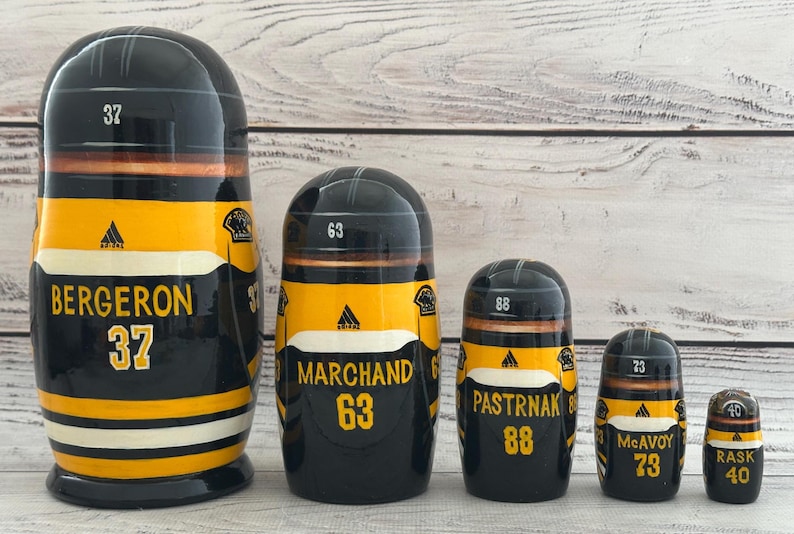May include: A set of five black, yellow, and white nesting dolls. Each doll features a hockey helmet design and the names and numbers of hockey players: Bergeron 37, Marchand 63, Pastrnak 88, McAvoy 73, and Rask 40.