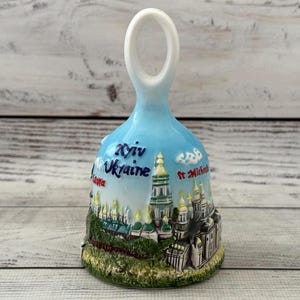 May include: A ceramic bell with a light blue sky background featuring a raised, colourful relief of the Kyiv Ukraine cityscape. The text Kyiv Ukraine and St. Michael are printed on the surface. The base is painted with green foliage details.