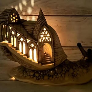 Puede incluir: Una escultura de caracol de cerámica con una iglesia gótica detallada e intrincada construida en su caparazón. La iglesia tiene ventanas y una puerta, y el interior está iluminado con una luz cálida y amarilla.