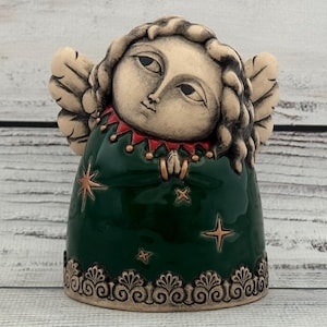 Op de afbeelding: Een keramieken engelenbeeldje met een donkergroene jurk en crèmekleurig gezicht en vleugels. De jurk van de engel is versierd met gouden sterren en een decoratieve rand. Het gezicht van de engel heeft een serene uitdrukking en de vleugels zijn gedetailleerd.