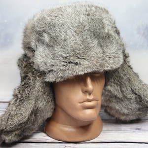 Puede incluir: Un gorro trapero de piel sintética gris con orejeras. El gorro está hecho de piel suave y mullida y tiene un estilo trapero clásico.