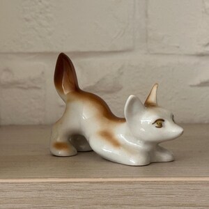 May include: A porcelain fox figurine in a playful pouncing pose. The fox is primarily white with smooth, glossy brown shading on its back, tail, and ears. It features painted gold-colored eyes and pointed ears, captured in a low, alert stance.