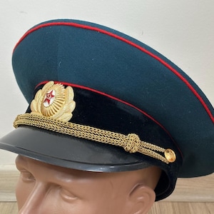 May include: A dark teal military hat with a red trim and a black visor. The hat features a gold emblem with a red star and gold rope detailing. The hat is a classic military style.