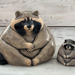 Peut inclure: Deux figurines de raton laveur en céramique en position assise avec des corps arrondis. Les deux personnages présentent une finition brillante, un pelage beige et gris, et les marques noires caractéristiques autour des yeux. Le plus grand est à gauche.