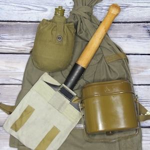 May include: A green canvas bag with a canteen, a shovel, and a metal cup. The bag has straps and buckles. The shovel has a wooden handle and a black metal blade. The canteen is green and has a brown strap. The cup is brown and has a metal handle.